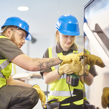 A female apprentice carpenter is installing handrails in a modern office refurbishment. She is being supervised by her co-worker. She is using a drill to make guide holes for the hand rails. They are wearing safety workwear.