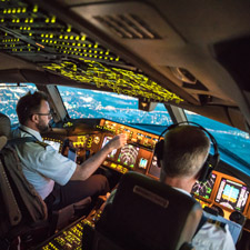 Two pilots sat in an aeroplane cockpit, operating illuminated buttons and looking out the windows to the sky ahead.
