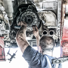 A mechanic stood underneath and working on an automotive engine.