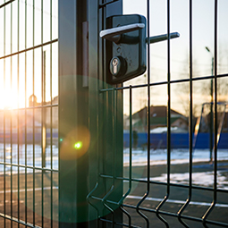 A close-up image of green mesh fencing, focused on the gate handle and lock. Behind the security fencing is a tennis court.