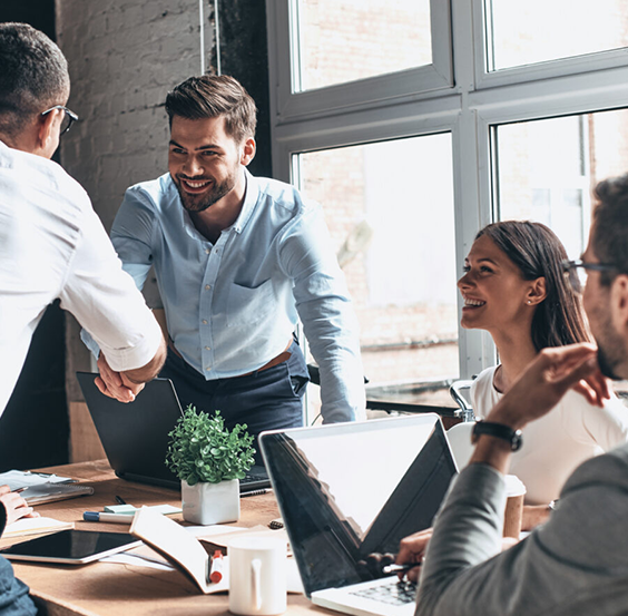 Two smiling men shaking hands over a desk, with a smiling man and woman also at their laptops alongside.